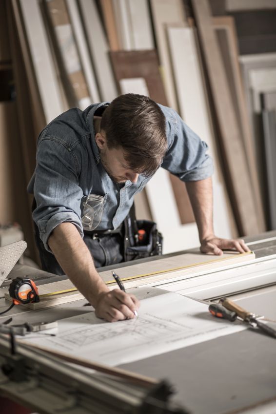 Carpenter measuring plans on a workbench, using tools in a woodworking shop.