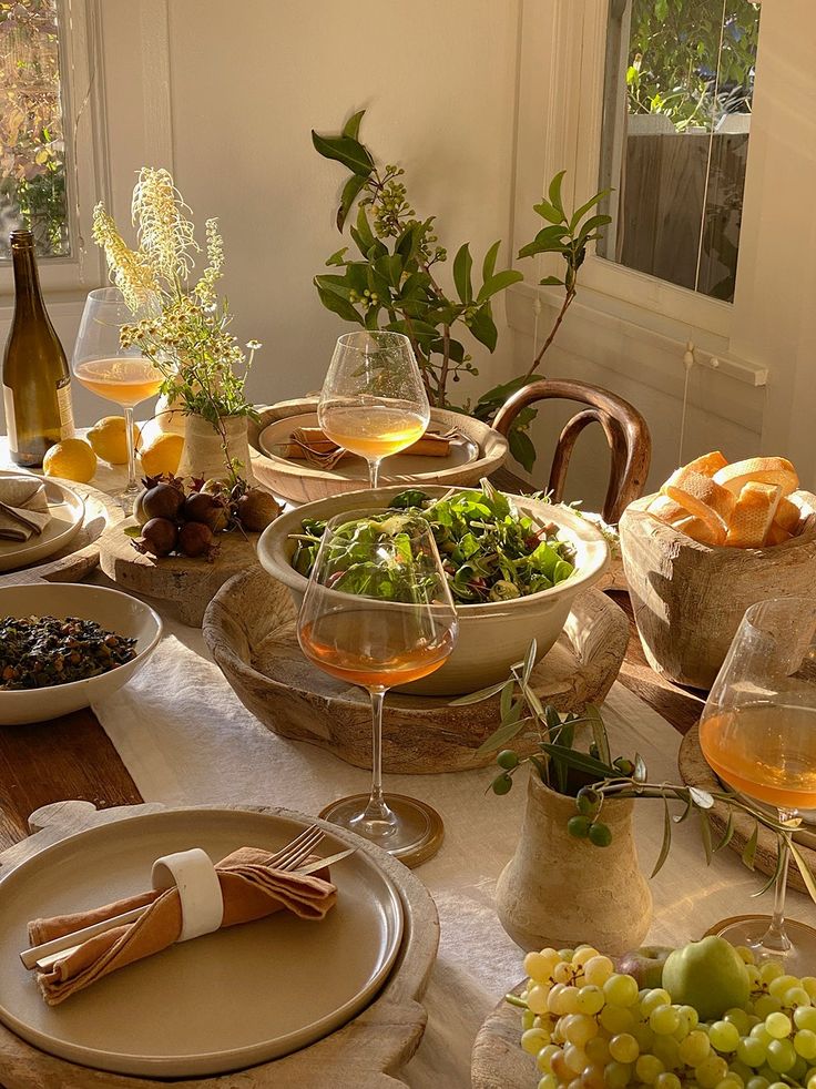 Elegant dining table set with fresh salad, fruits, and wine in sunlit room with rustic decor and greenery.