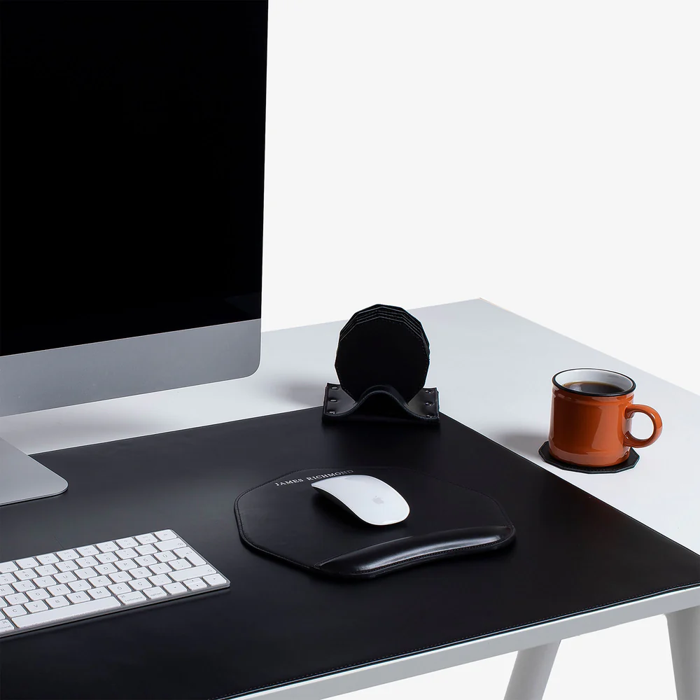 Modern workspace with computer, keyboard, mouse on pad, coasters, and coffee mug. Minimalist black and white desk setup.