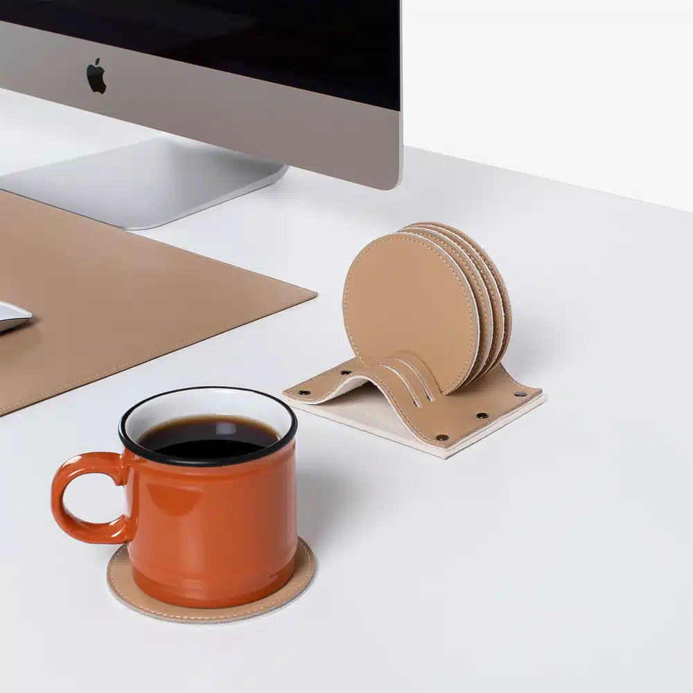 Stylish desk setup with computer, orange mug, and beige leather coasters on a modern white surface.