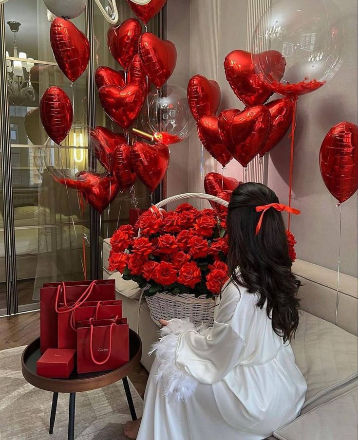 Woman in white seated with red roses, heart balloons, and red gift bags for a romantic celebration.