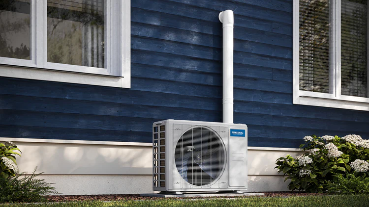 Outdoor heat pump unit installed on a house exterior wall, surrounded by plants and a blue wooden facade.