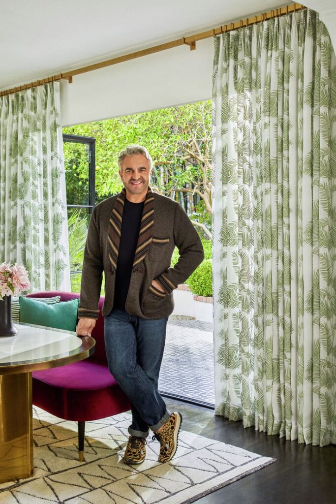 Man in stylish outfit stands by window with leafy curtains, modern interior design.