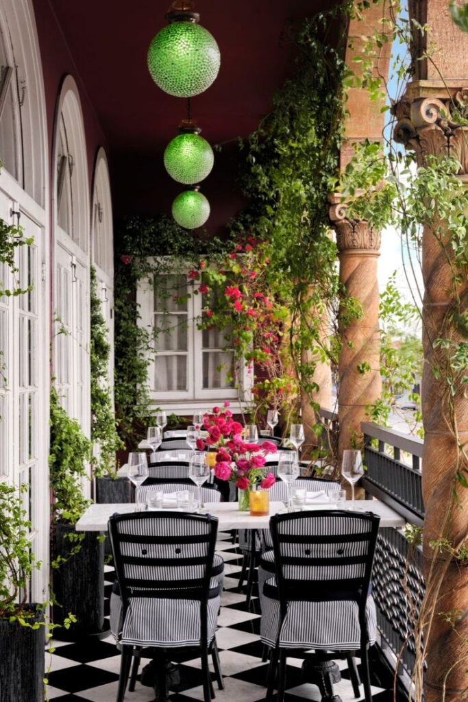 Charming outdoor dining with floral decor, black chairs, green lanterns, and lush vines on a checkered floor veranda.