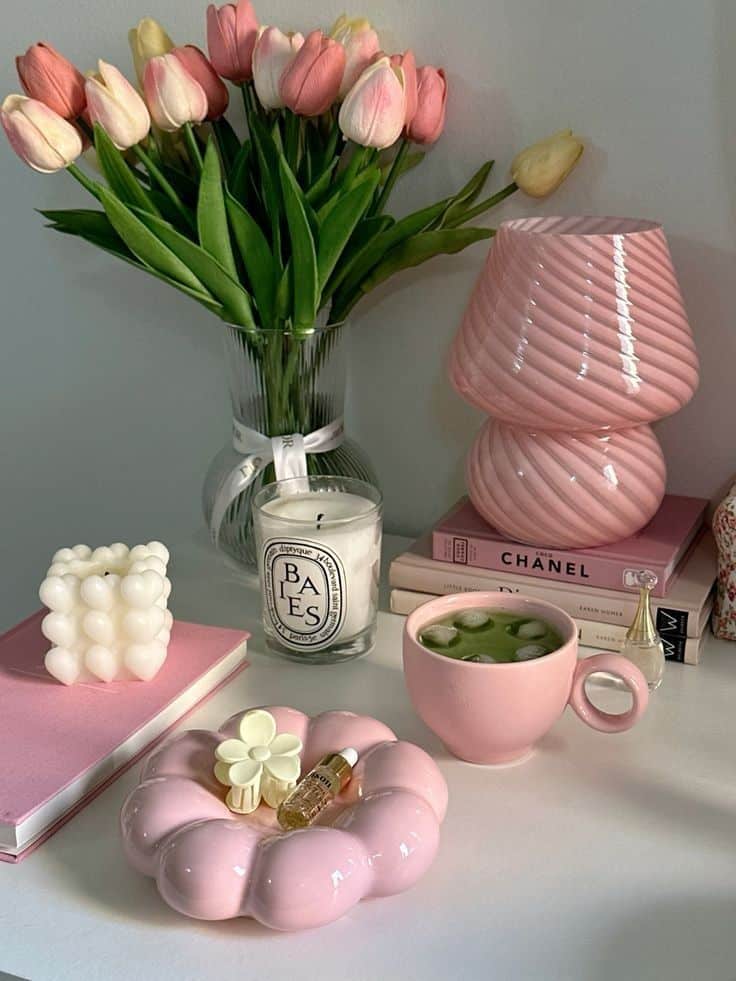 Pink tulips in a vase with a pink lamp, books, and decor on a desk; chic and cozy home aesthetic.