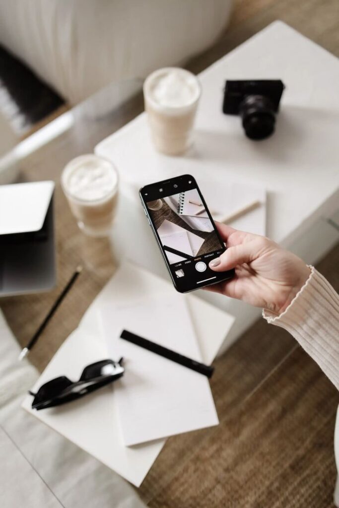 Person taking a photo of a workspace with smartphone, coffee, camera, and notepad. Cozy and creative setup.