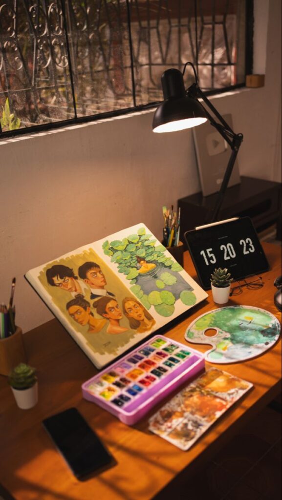 Artist's workspace with sketchbook, watercolor paints, and digital clock on wooden desk.