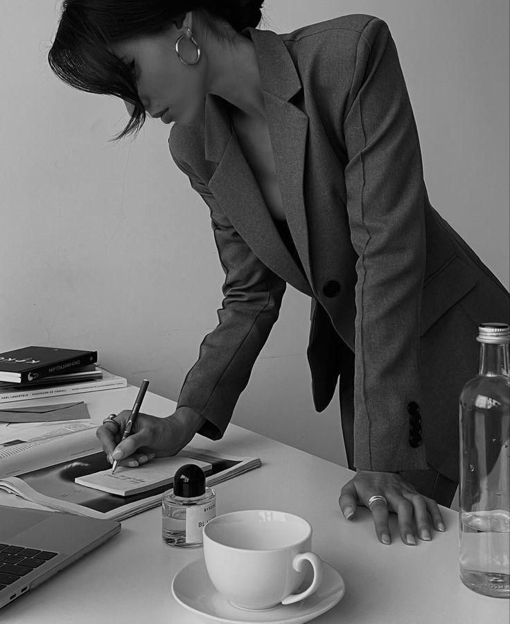 Businesswoman in a grey suit writing at a desk with books, laptop, perfume, and coffee cup. Black and white photo.