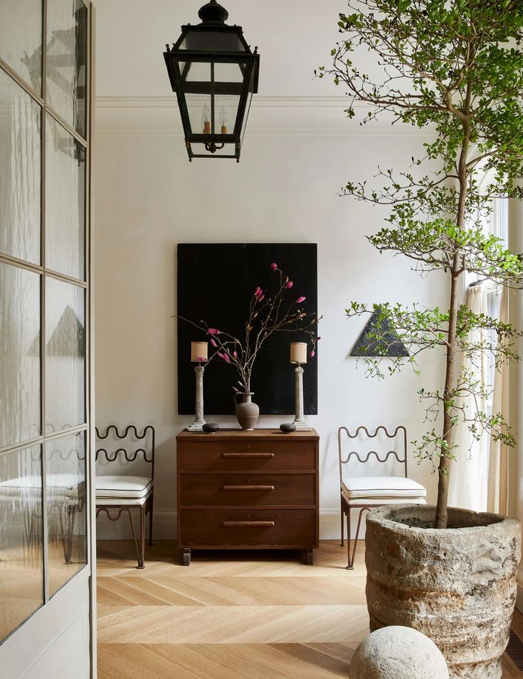 Elegant hallway with a wooden dresser, modern chairs, large potted plant, and stylish decor in a minimalistic setting.