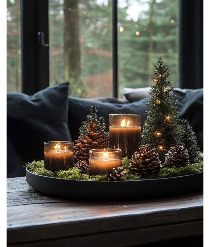 Cozy winter decor: candles, miniature trees, and pinecones on a tray by a window, creating a warm holiday ambiance.