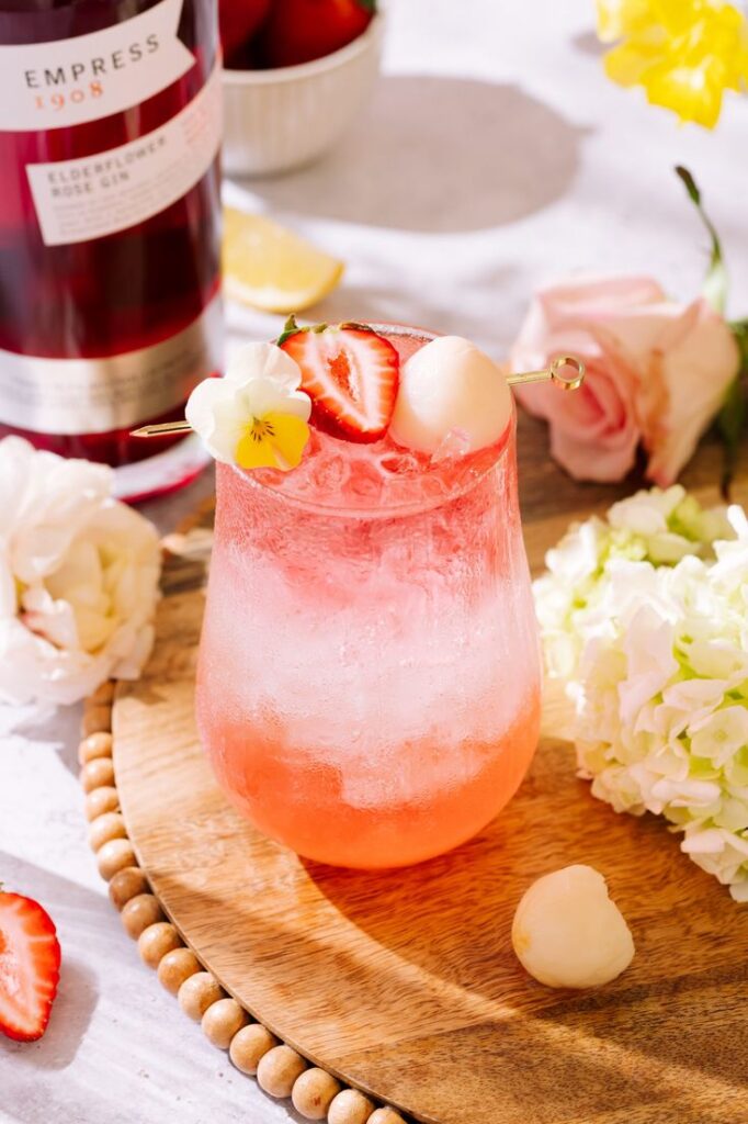 Refreshing pink cocktail with strawberries, lychee, and flowers, served in a glass with ice on a wooden tray.