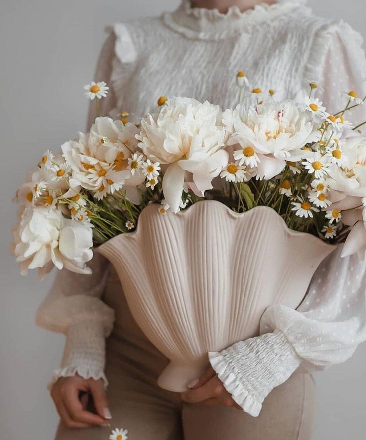 Person holding a vase with white peonies and daisies, wearing a white blouse. Elegant floral arrangement display.