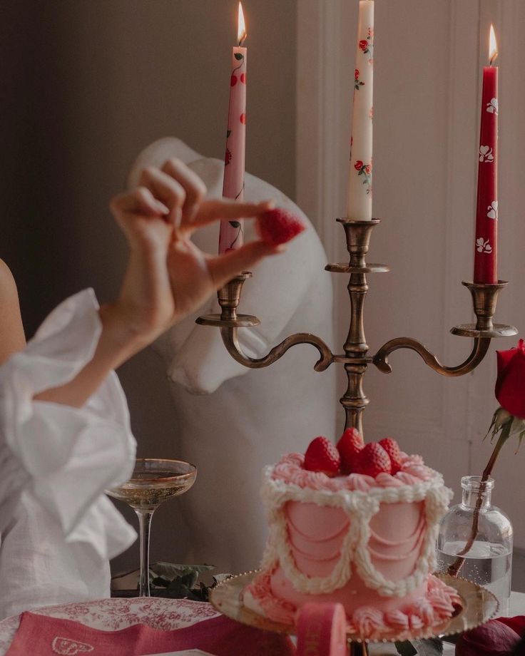 Elegant pink cake with raspberries, lit candles, and a romantic setting with a hand holding a strawberry.