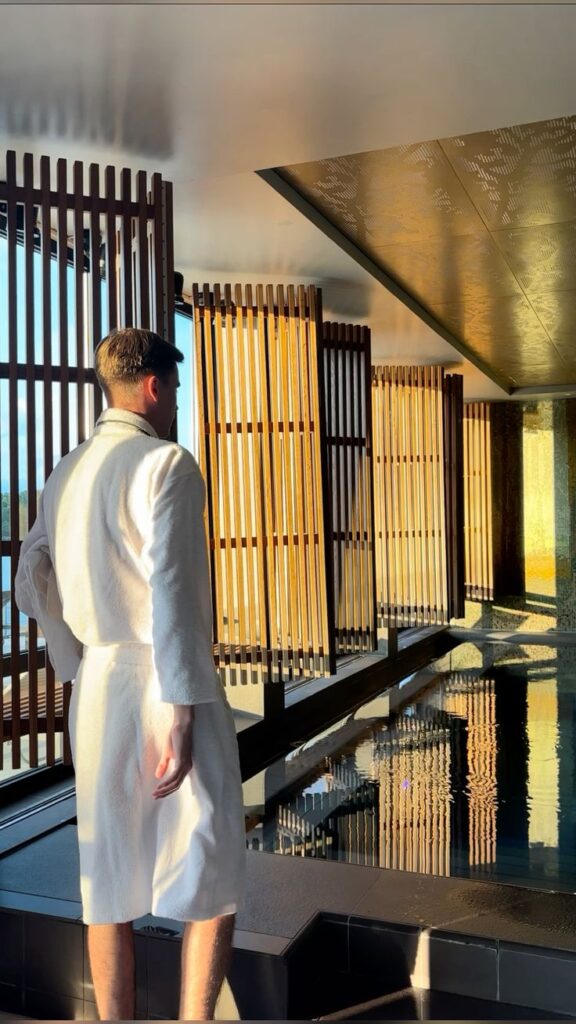 Person in a robe stands by a serene indoor pool with wooden decor, bathed in warm light.