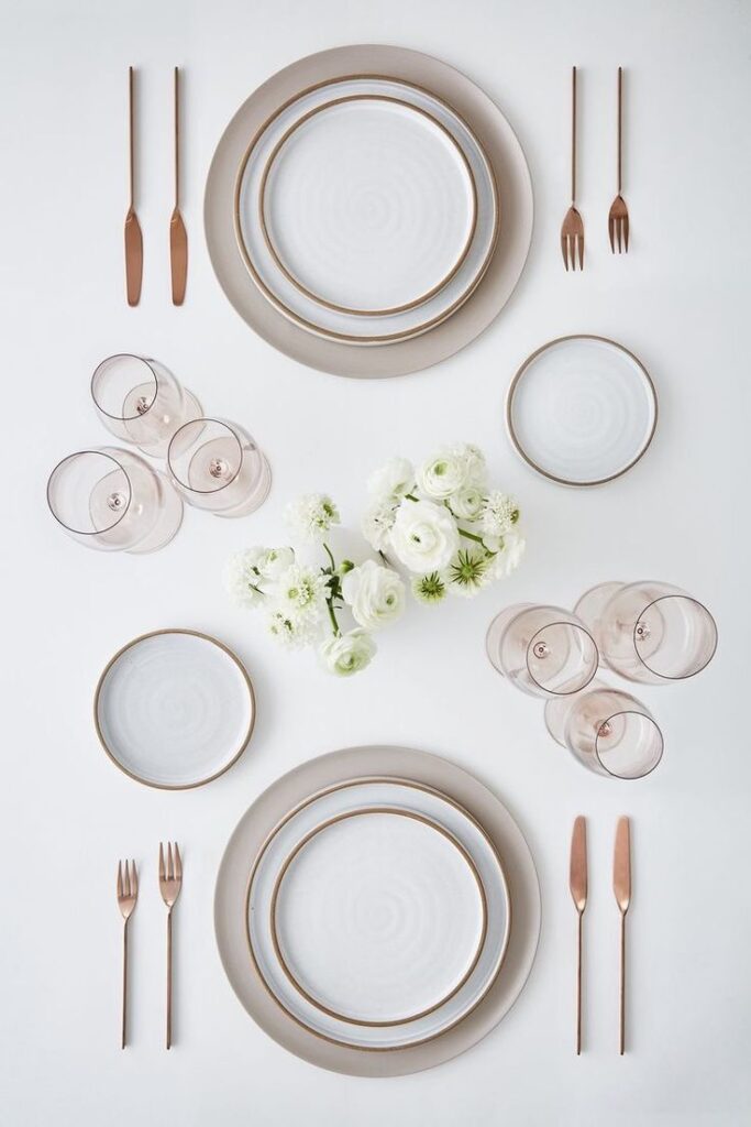 Elegant table setting with white plates, rose gold cutlery, blush glassware, and white flowers on a light background.