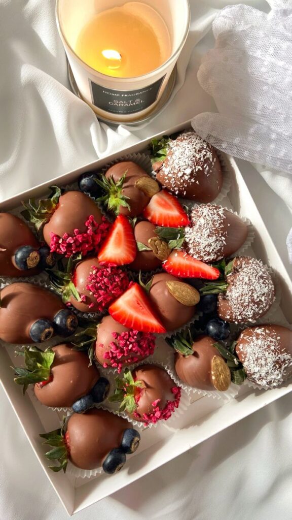 Box of assorted chocolate-covered strawberries with toppings next to a lit salted caramel candle.