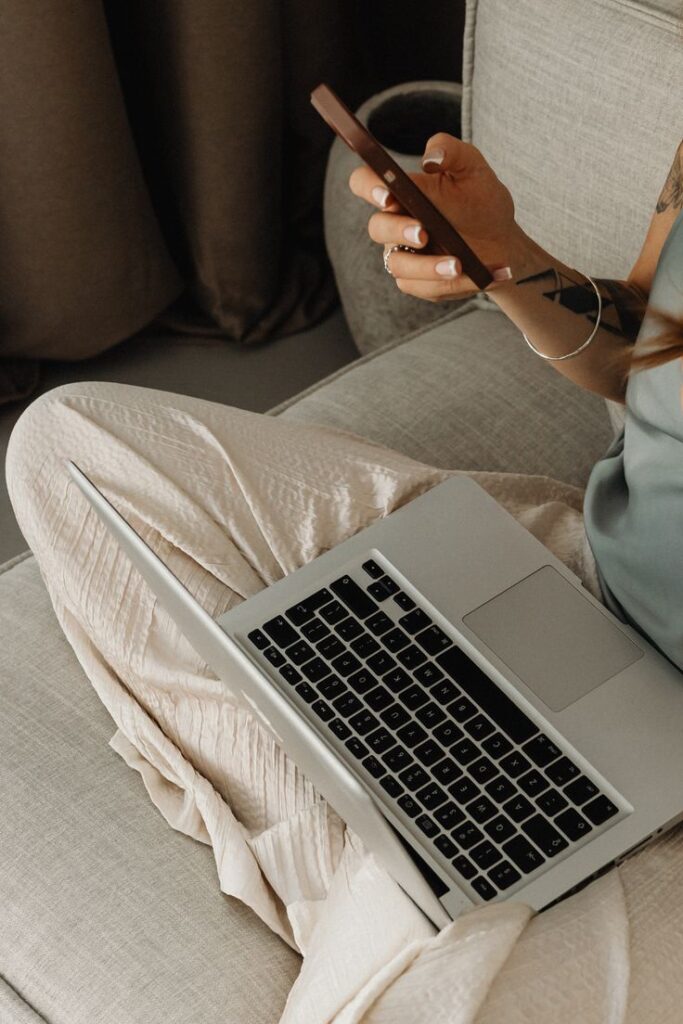 Person multitasking with a smartphone and laptop on a cozy sofa, showcasing remote work flexibility.