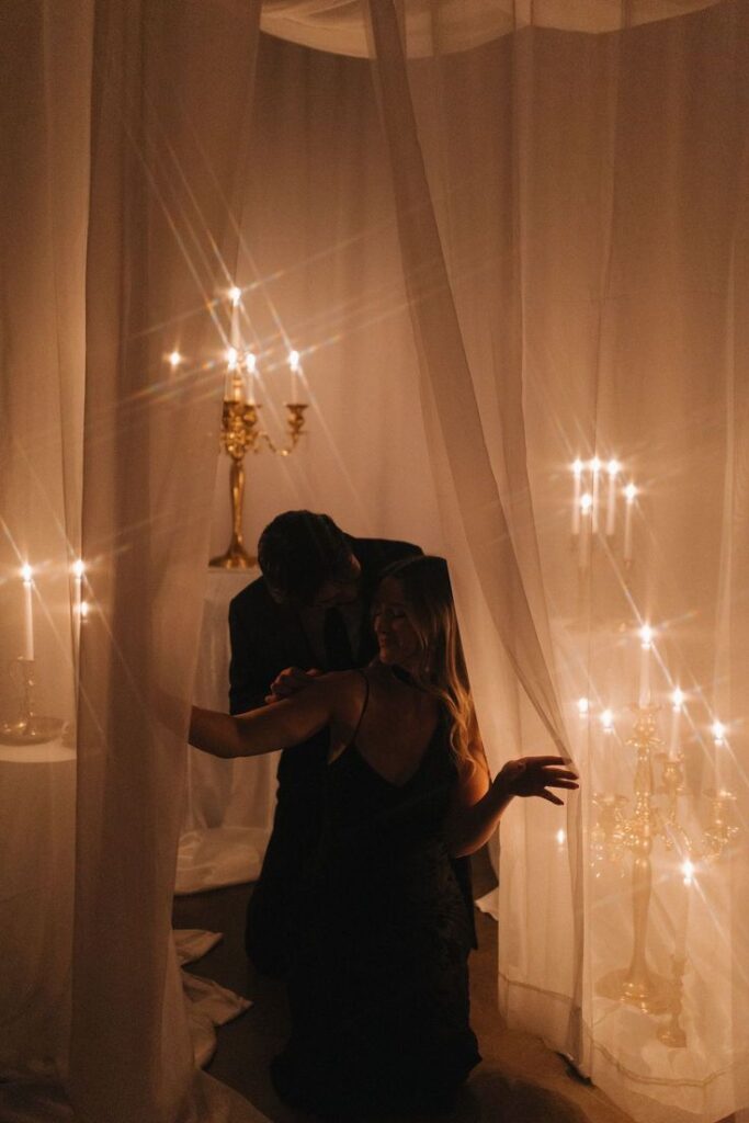 Romantic couple illuminated by candlelight, surrounded by sheer curtains and golden candelabras.