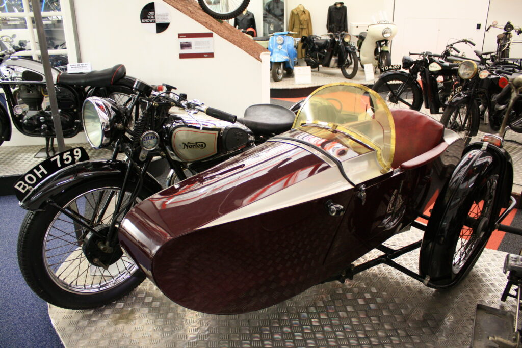 Vintage Norton motorcycle with a sidecar on display in a museum showcase of classic vehicles.