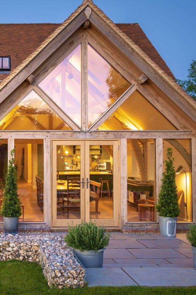 Cozy timber-framed home with large glass windows, warm interior lighting, and outdoor greenery at dusk.