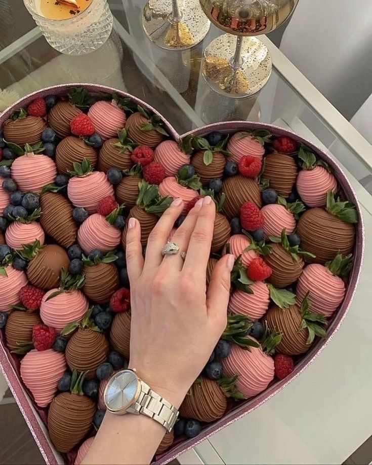 Heart-shaped box of chocolate-covered strawberries, raspberries, and blueberries with a hand wearing a watch.