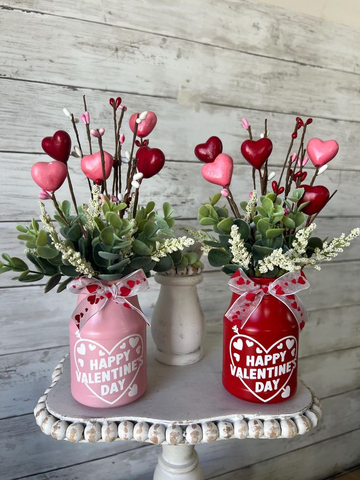 Valentine's Day heart decorations in pink and red jars with greenery on a rustic table.