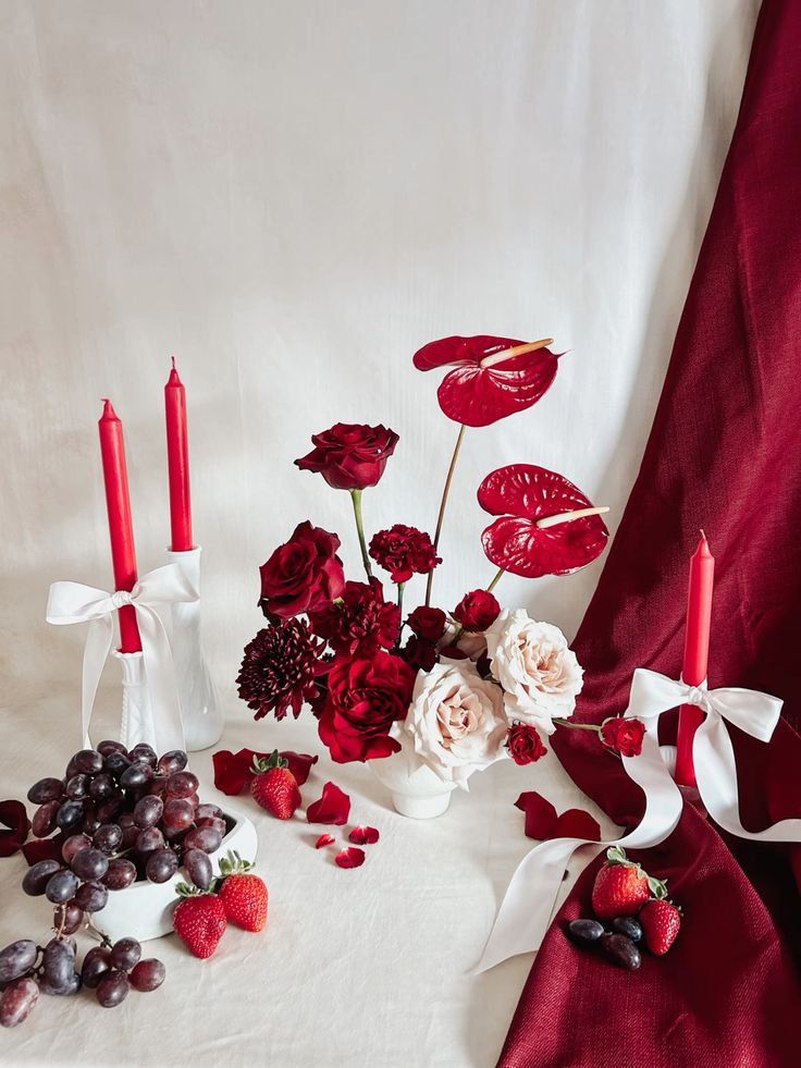 Elegant red roses, candles, and fruit arrangement with white fabric, creating a romantic floral decor setting.