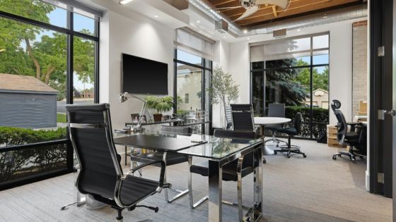 Modern office with glass desks and black chairs, large windows, natural light, and greenery.