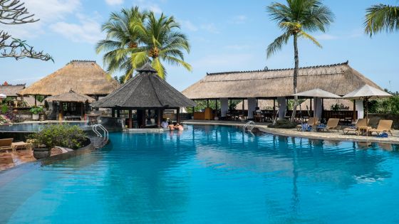 Tropical resort pool with palm trees and thatched-roof cabanas under a clear blue sky. Relaxing vacation destination.