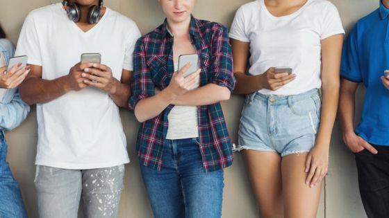 Group of diverse friends using smartphones while standing against wall, showcasing modern digital lifestyle.