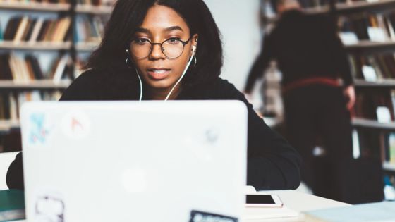 Young woman with glasses studying on a laptop in a library, wearing earphones.