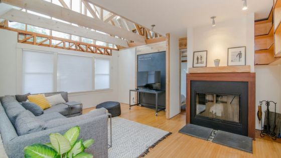 Modern living room with large gray sofa, fireplace, and natural light from windows and exposed beams.