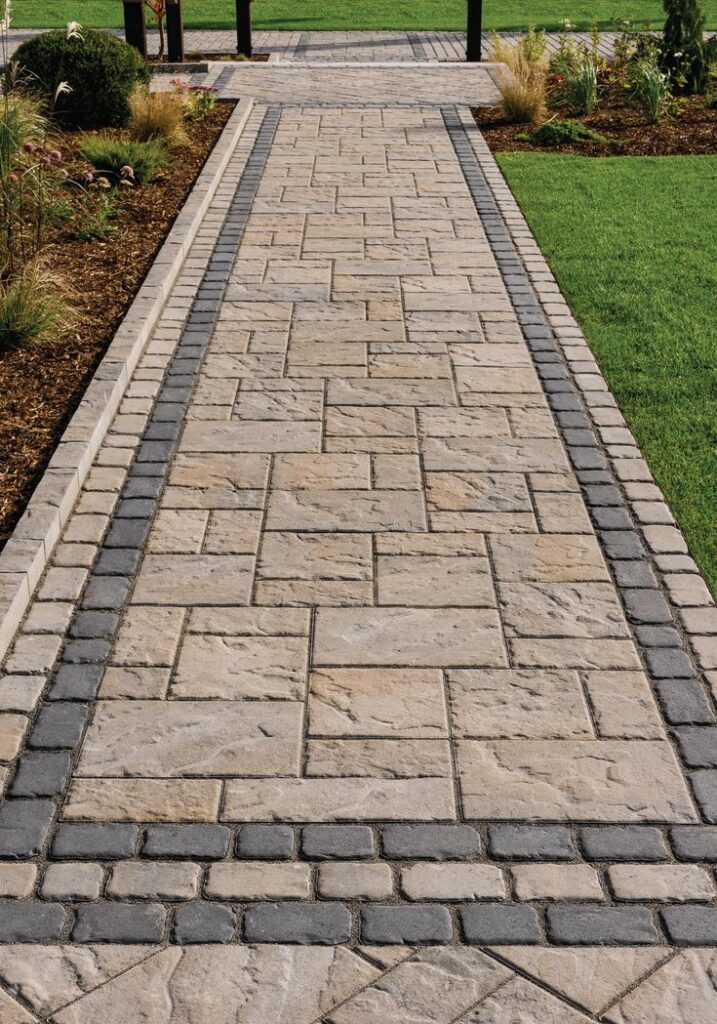 Stone pathway with geometric pattern set in garden, bordered by grass and plants, leading to outdoor structure.