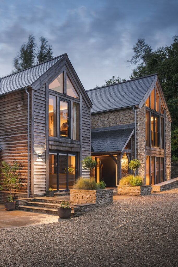 Modern rustic house exterior with large glass windows, stone and wood design, surrounded by trees at dusk.