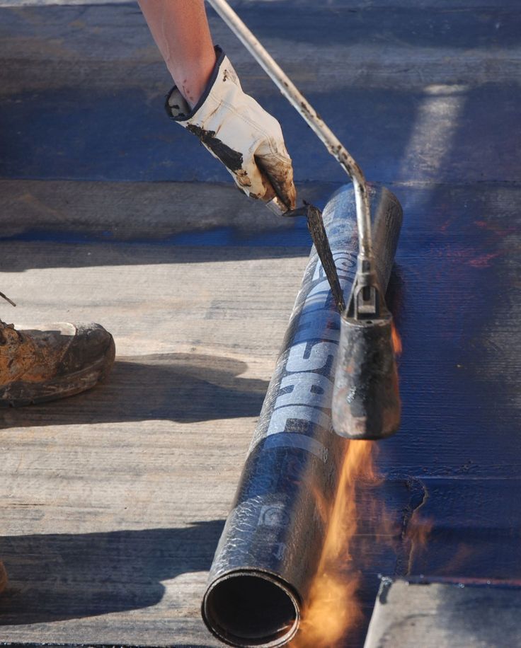 Worker applying torch-down roofing membrane with a propane torch, showcasing construction work.