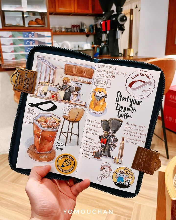 Hand holding an illustrated coffee journal in a cozy café, featuring sketches and motivational coffee quotes.