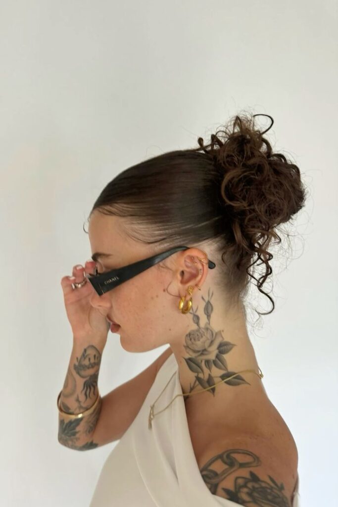 Woman with updo and floral tattoos wears sunglasses and gold earrings in a minimalist white top.