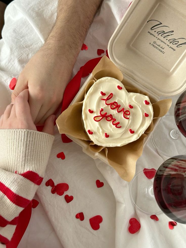 Romantic setting with heart-shaped cake and love you icing, surrounded by red hearts and holding hands.