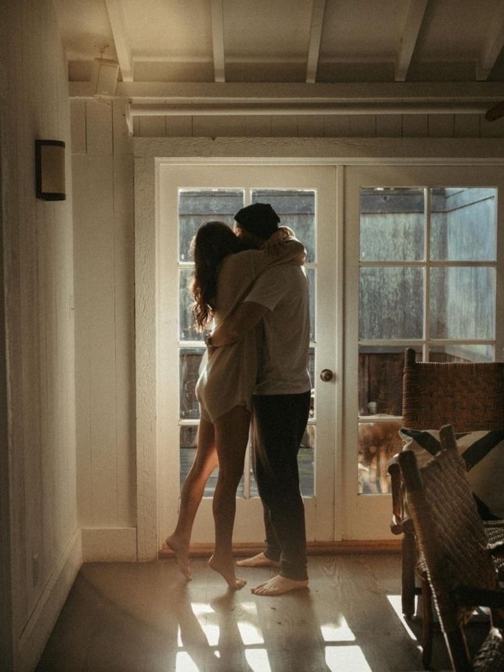 Couple hugging warmly in sunlit room, standing near French doors. Cozy and intimate home setting.