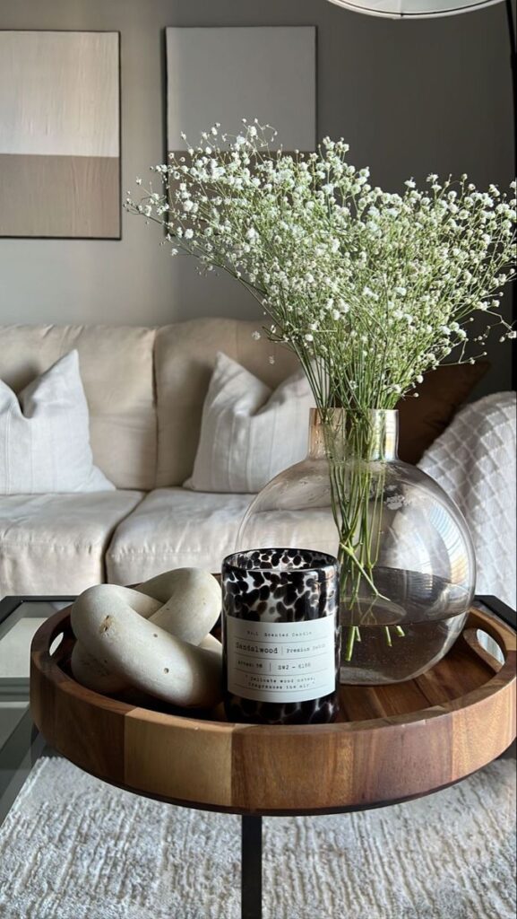 Elegant living room decor with vase of flowers, scented candle, and modern wooden tray on a glass coffee table.