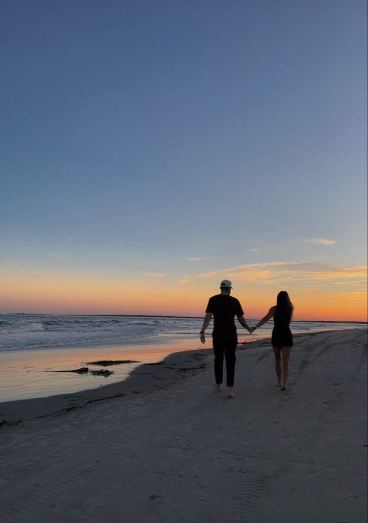 Couple holding hands while walking along a beach at sunset, serene ocean waves under a colorful sky.