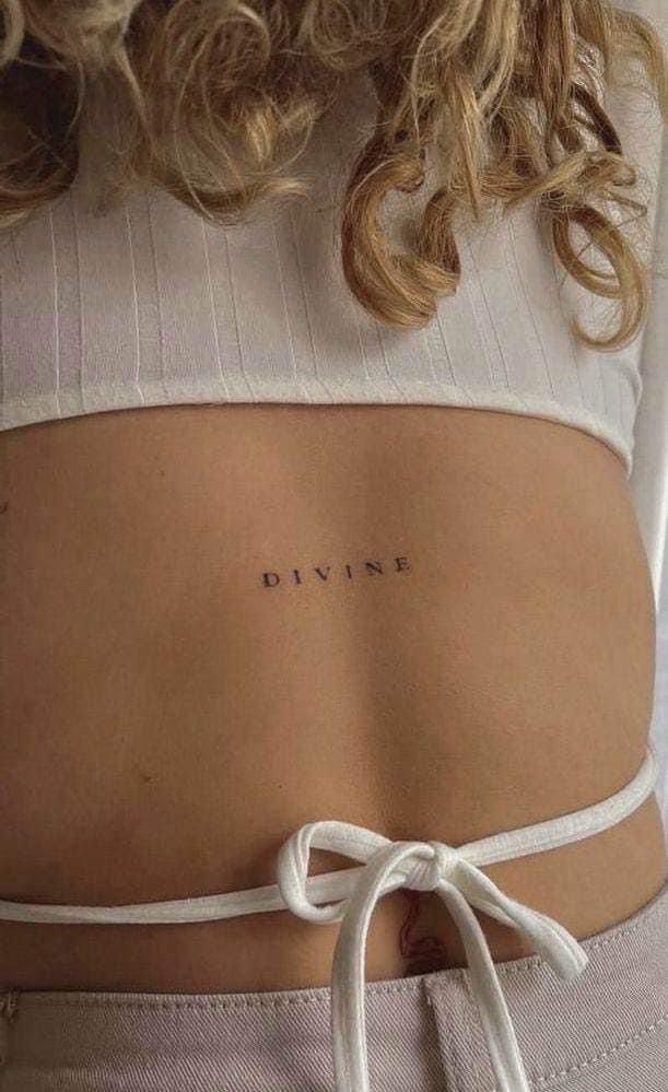 Lower back Divine tattoo, woman wearing a white crop top with tie-back detail, curly blonde hair.