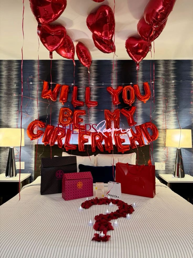 Romantic proposal setup with red balloons, gifts on a bed, and heart-shaped decor asking Will you be my girlfriend?