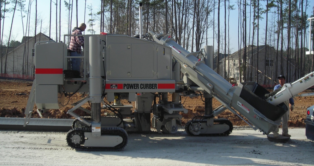 Road construction with a Power Curber machine laying concrete in a forested area.