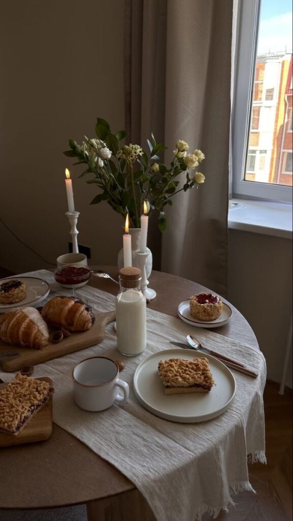 Cozy breakfast setting with croissants, cakes, and milk on a table near a window, decorated with flowers and candles.