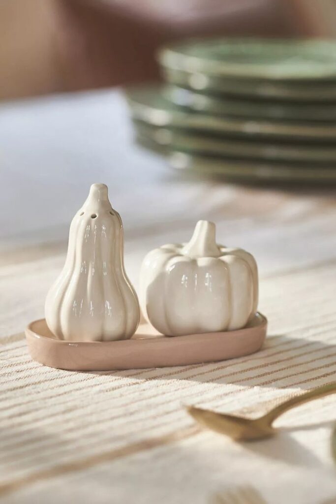 Ceramic pumpkin and gourd salt and pepper shakers on a striped tablecloth, with stacked green plates in the background.