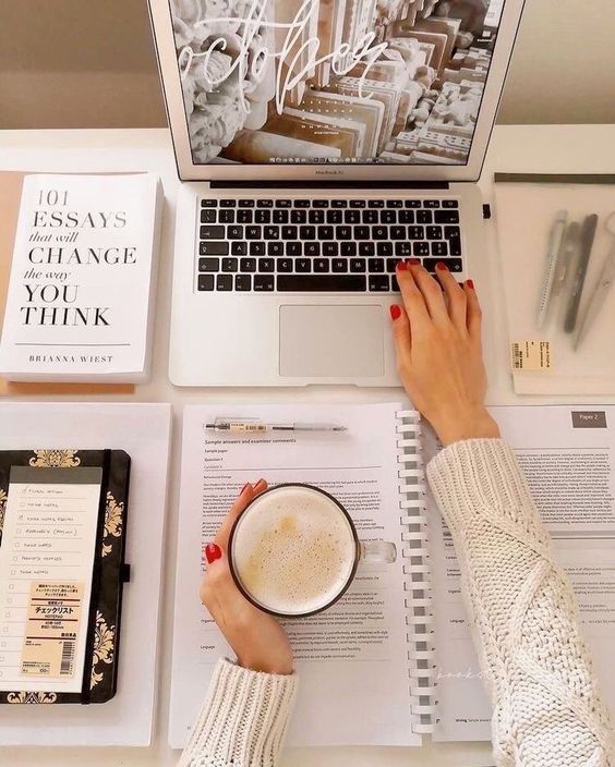 Woman working at a cozy desk with a laptop, coffee, and a book titled 101 Essays That Will Change the Way You Think.