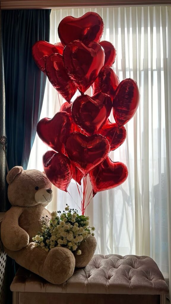 Teddy bear with white roses and heart-shaped red balloons by a window, creating a romantic Valentine’s Day setting.