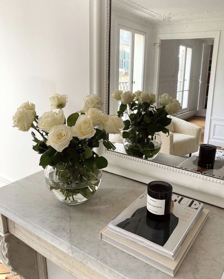 Elegant white roses in a glass vase on a marble table, reflected in a mirror, with a candle and magazine.