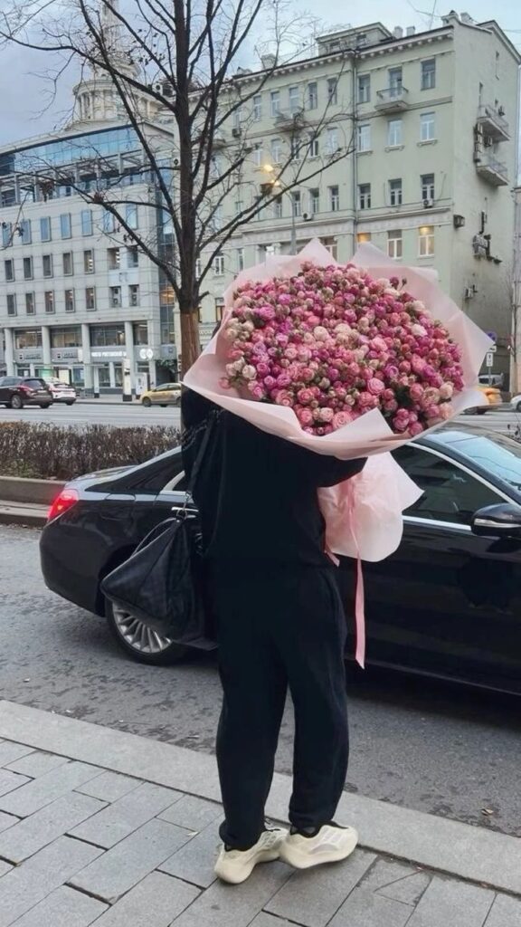 Person holding a large bouquet of pink flowers on a city street, standing beside a black car.